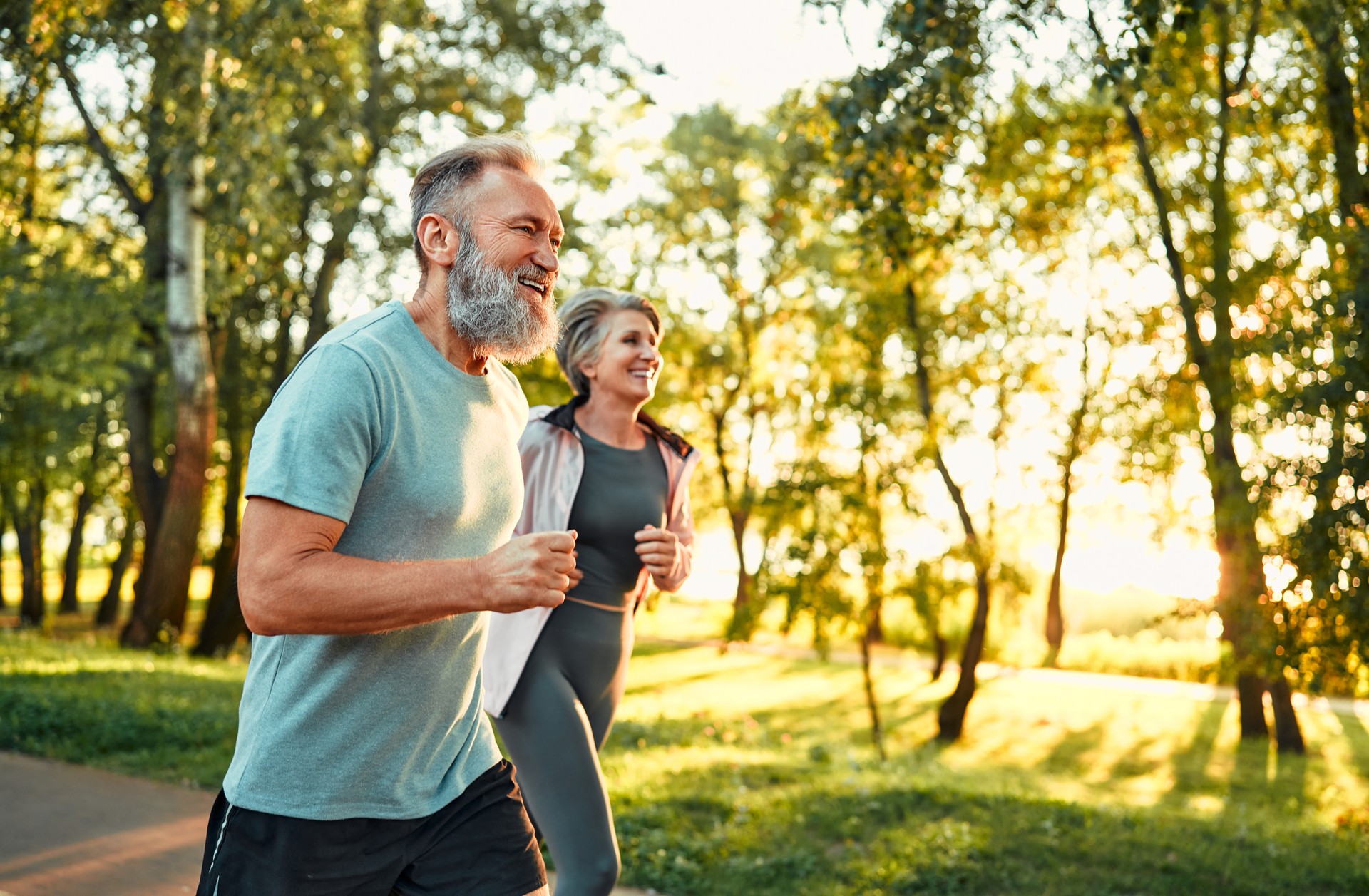 Cardio training outdoors. Side view of caucasian grey bearded man and beautiful old woman running along summer park with sunlight on background. Cheerful retired people leading active lifestyle.